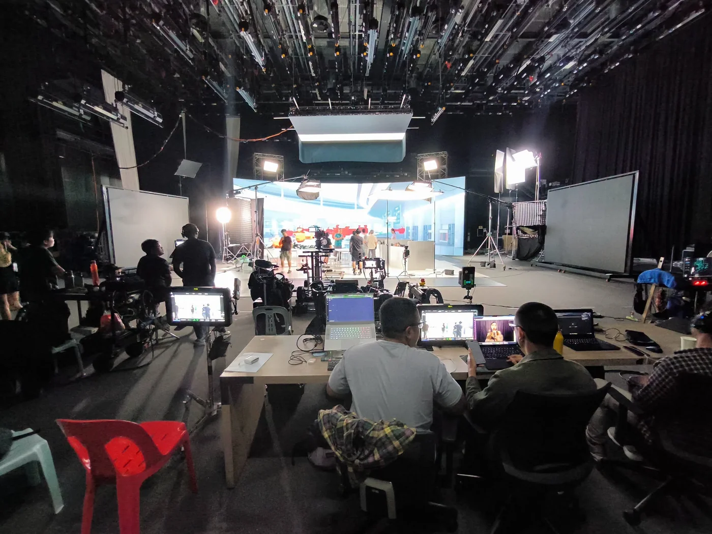 Crew, cameras, practical tables and red stools share the studio floor in front of a virtual environment.