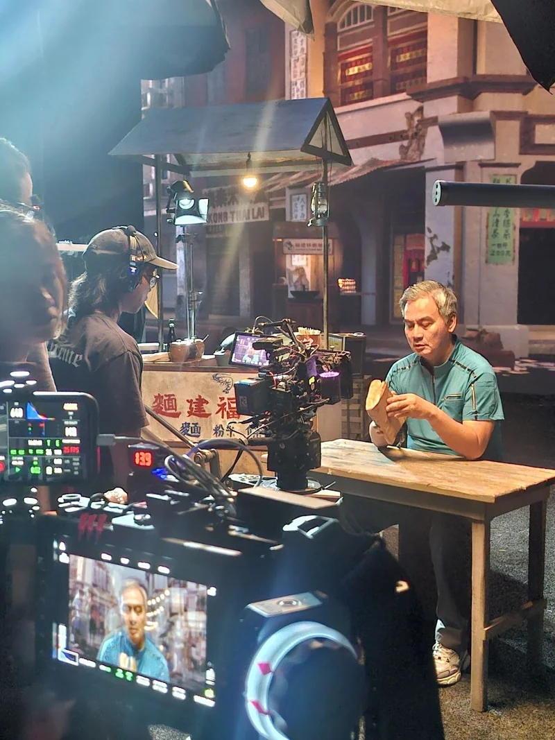 The actor holds the recipe book at a practical table while the camera and crew prepare the shot.