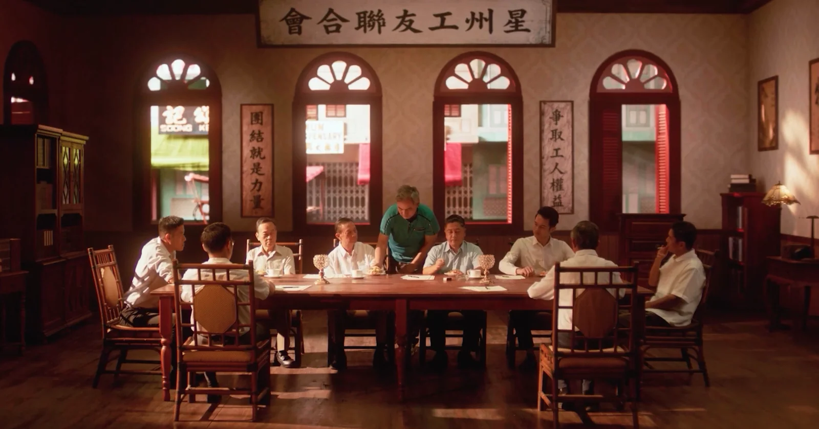 People sit around a long wooden table in the period Union Hall scene.